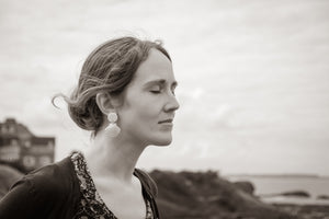 Model with handmade polymer clay seashell earrings pictured by the coast, with eyes closed and hair blowing in the wind.